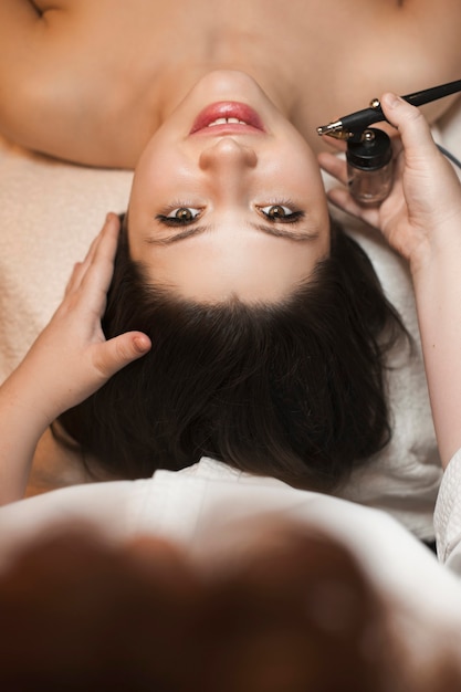 Upper view of a cute young woman looking at camera while having oxygen procedure in a spa salon by a cosmetologist.