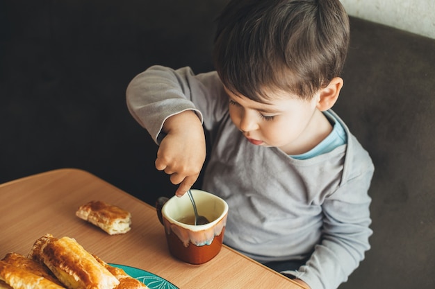 Foto di vista superiore di un ragazzo caucasico seduto al tavolo e bere un tè con un cucchiaio mentre mangia dei biscotti