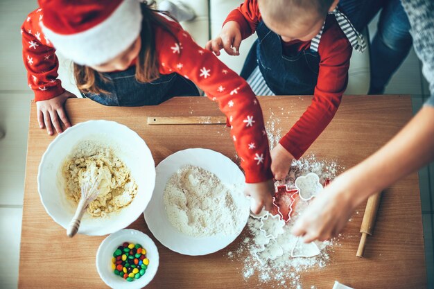 サンタの服を着てクリスマスに小麦粉と生地を使用してクッキーを作る子供たちの上面写真