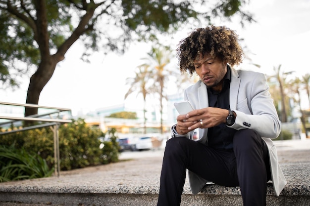 Photo upset african american middle aged male entrepreneur sitting on stairs outdoors and using cellphone