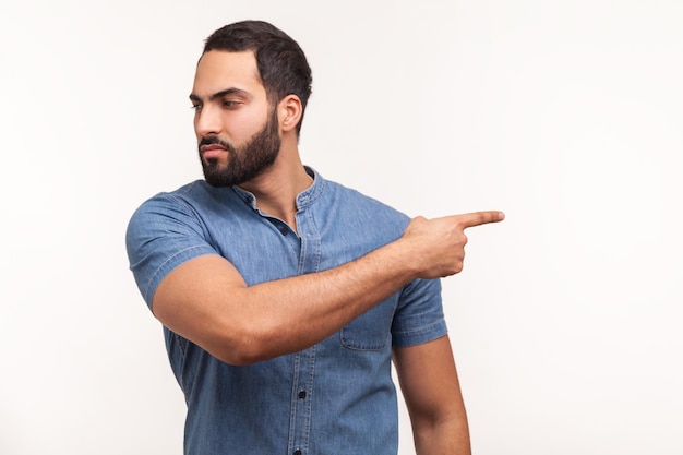 Uomo arrabbiato sconvolto con la barba in camicia blu che punta il dito lontano guardando con espressione risentita confusa con il conflitto che calcia fuori studio indoor girato isolato su sfondo bianco