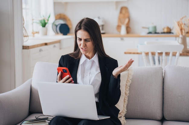 Foto sconvolta la giovane imprenditrice arrabbiata seduta sul divano di casa in abito nero con il portatile sconvolto dalle cattive notizie