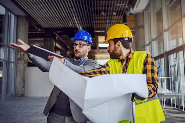 Upset architect arguing with construction worker and showing him his mistake. Worker holding blueprints and defending himself. Building in construction process interior.