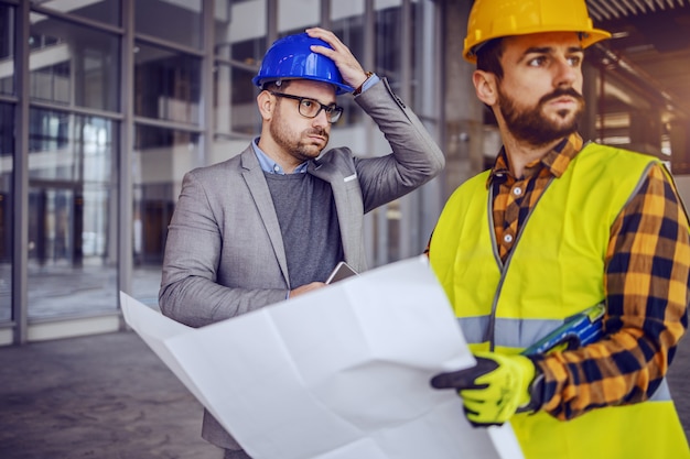 Upset architect holding his head and thinking about mistake he made on blueprints. construction worker holding blueprints and looking away.