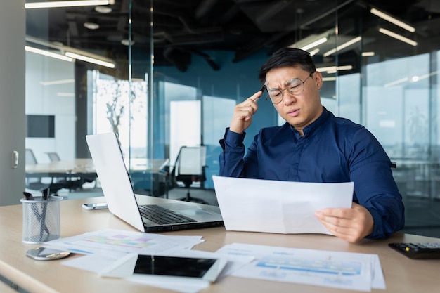 Расстроенный азиатский бизнесмен за бумажной работой внутри современного офисного финансиста-мужчины, работающего с ноутбуком