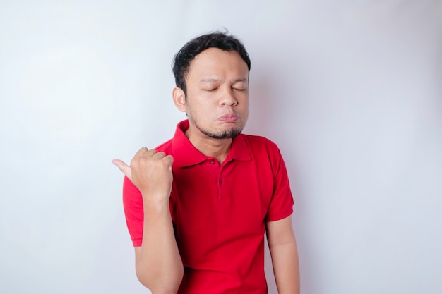 Uomo asiatico sconvolto che indossa una maglietta rossa che punta allo spazio della copia accanto a lui isolato da uno sfondo bianco