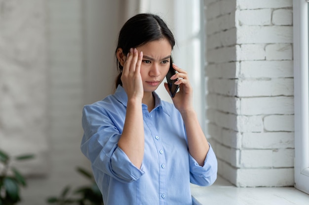 Upset asian woman standing by window having phone conversation