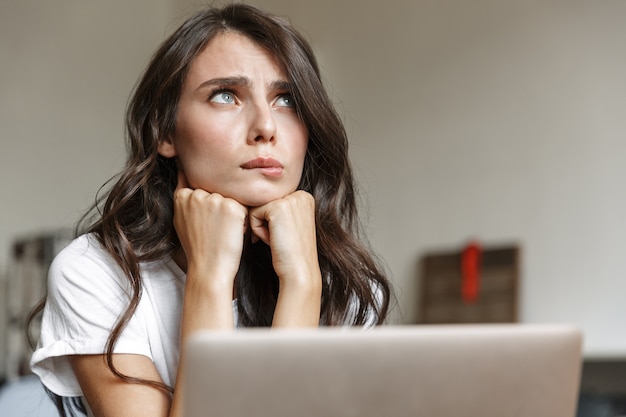 Foto donna attraente sconvolta con i capelli lunghi ricci che guarda in alto e usa il laptop mentre è seduta al tavolo in camera