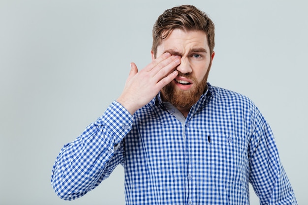 Upset bearded young man standing and rubbing his eye over white wall