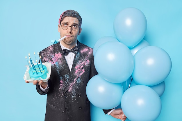 L'uomo di compleanno sconvolto soffia un fischio tiene un mazzo di palloncini gonfiati e una torta dolce con candele burnnig si sente annoiato sulla festa vestita formalmente isolata su sfondo blu evento festivo della gente