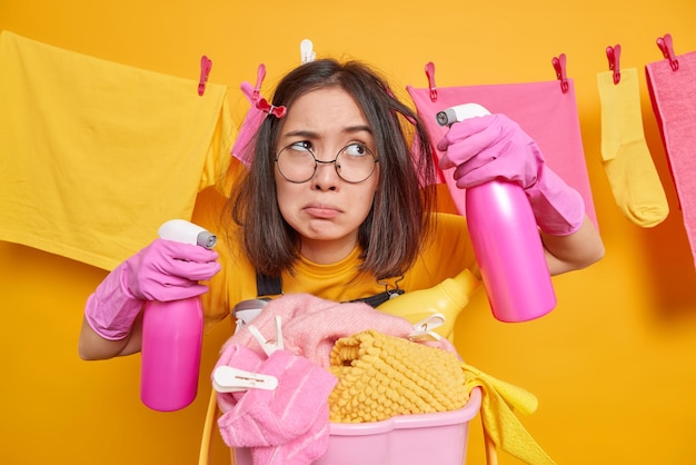 Upset bothered asian housewife holds two bottles of cleaning\
detergent looks aside wears round spectacles poses near basket of\
dirty laundry poses against yellow background with clothesline\
behind