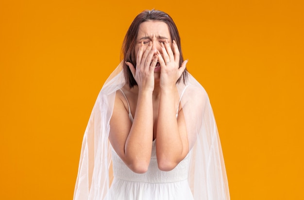 Upset bride in beautiful wedding dress crying hard covering eyes with palms standing over orange wall