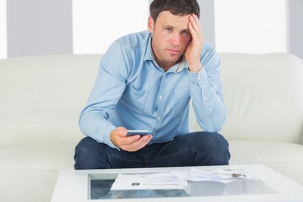 Upset casual man using calculator and looking at camera paying bills