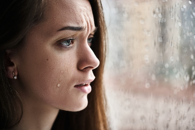 Foto sconvolto pianto donna con lacrime agli occhi che soffrono di shock emotivo, perdita, dolore, problemi di vita e relazione di rottura vicino alla finestra con gocce di pioggia. la femmina ha ricevuto cattive notizie