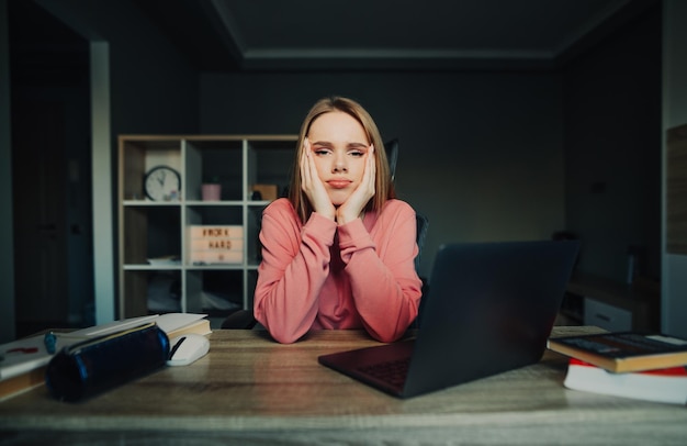 upset cute female student sitting at desk at home with laptop and books and looking at camera