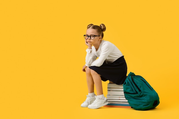 Расстроенная милая школьница сидит на куче книг на желтом фоне студии