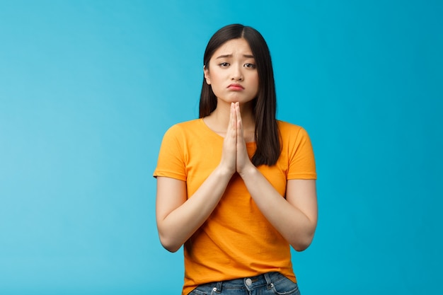 Upset cute silly asian girl praying, plead for help, pouting frowning need, make pitty face, hold hands pray begging for favor, apologizing feelings guilty sad, stand blue background