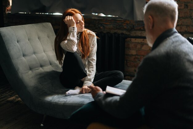 Upset depressed redhaired young woman talking about her problems at psychologist appointment Unrecognizable psychotherapist listening patient and giving advices Concept of psychological treatment