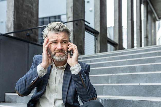 Upset and disappointed mature businessman sitting on stairs of office building outside man in