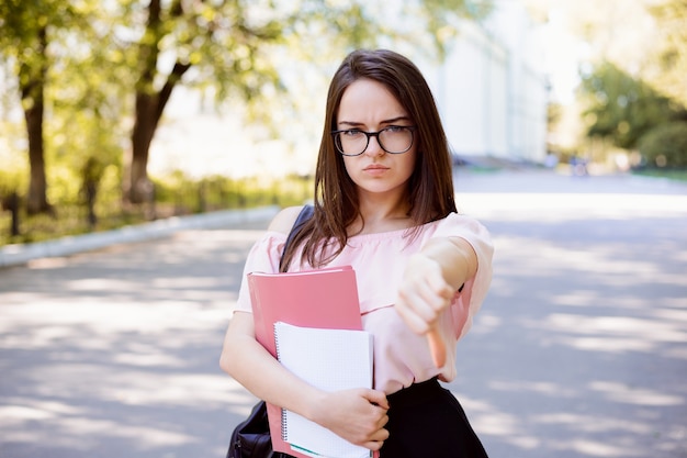 屋外に立って親指を下に見せている動揺して失望した若い学生の女の子