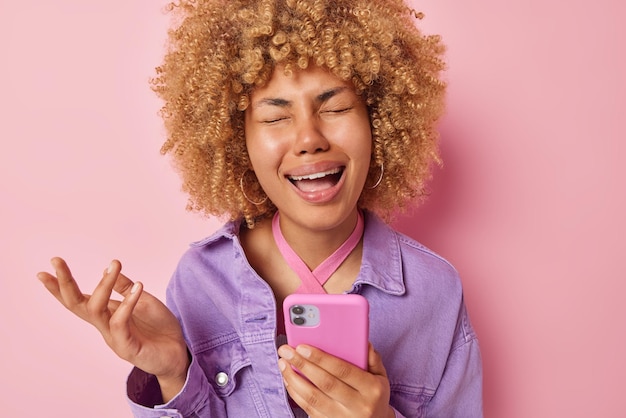 Upset dissatisfied woman with curly hair holds mobile phone\
keeps hand raised expresses negative emotions has doleful\
expression sad as didnt receive call from boyfriend dressed in\
purple jacket