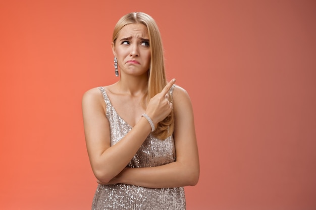 Upset doubtful unsure cute blond funny glamour girl in silver evening dress cringing grimacing hesitant pointing looking upper right corner suspicious, uncertain standing red background.