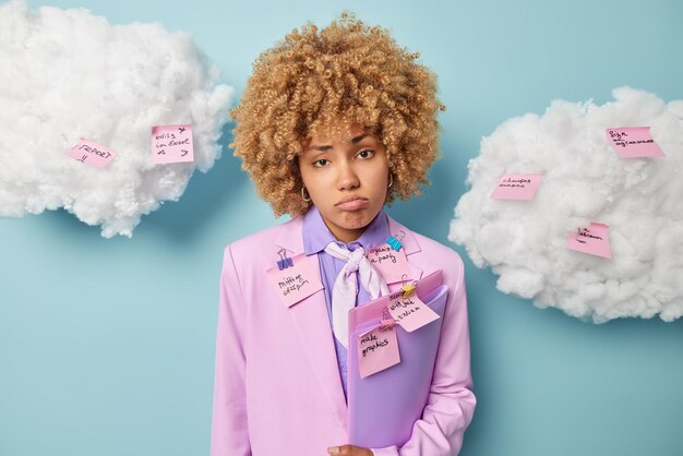 Upset female office worker dressed formally carries folders\
wrote ideas and tasks to do on sticky notes feels exhausted poses\
against blue background white clouds around student busy preparing\
report