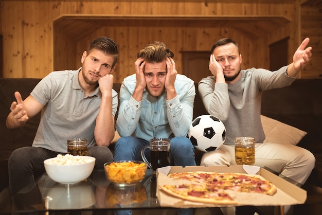 The upset friends watch a football near the table with a food