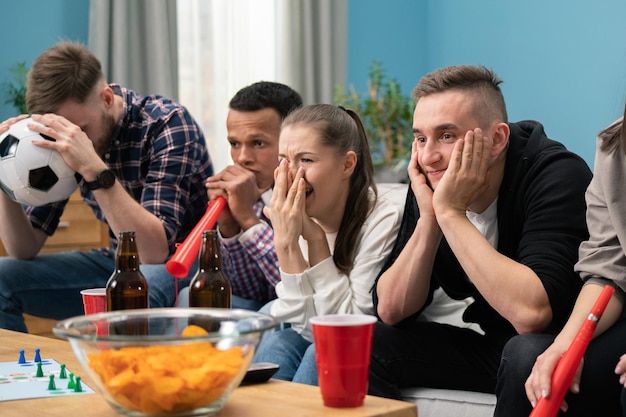 Расстроенные друзья смотрят футбол и пьют дома, сидят на диване, студенты смотрят спорт