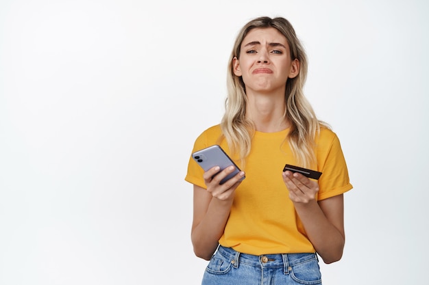 Фото Расстроенная девушка держит смартфон и кредитную карту, концепция женщины без денег, не может что-то купить, стоя на белом