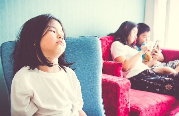 Upset girl sitting on chair Mother enjoying with brother on sofa at home