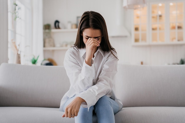 Upset girl sitting on sofa dressed in white shirt blue jeans crying leans on hand at kitchen