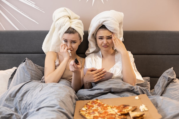 Расстроенные девушки лежат на кровати, смотрят телевизор или фильм и плачут. молодые европейские женщины с обернутыми полотенцами на головах. концепция вечеринки девочек дома. интерьер спальни в современной квартире