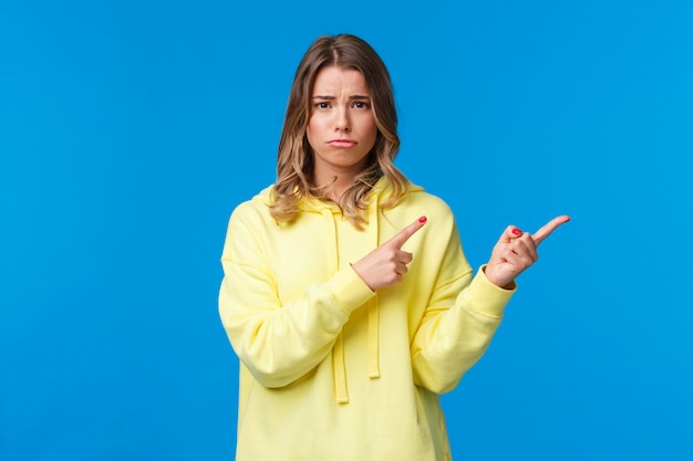 Upset and gloomy cute blond teenage girl frowning and sulking from unfair situation, someone offended her, feel jealous or regret, frowning camera and pointing fingers right, 