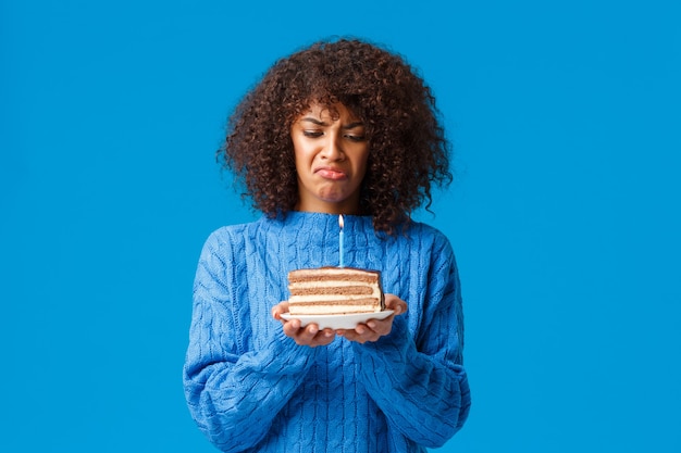 Upset and gloomy, distressed young african-american woman hate celebrating birthday feeling older, looking bothered and displeased at b-day cake with lit candle