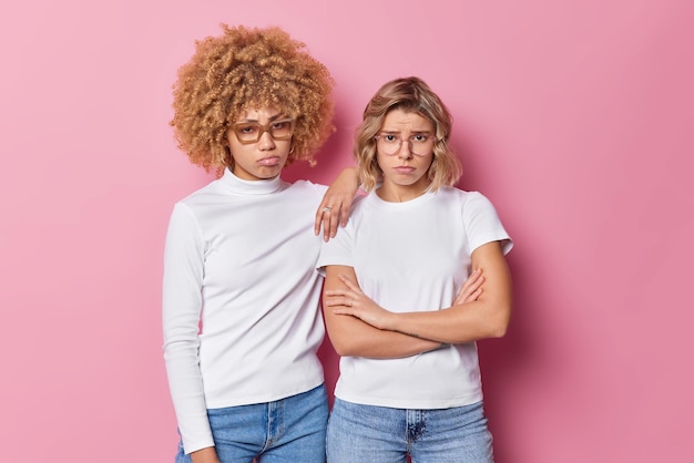 Upset gloomy female friends look frustrated have grumpy expressions wear casual clothes express negative emotions isolated over pink background Worried discontent young women have problems