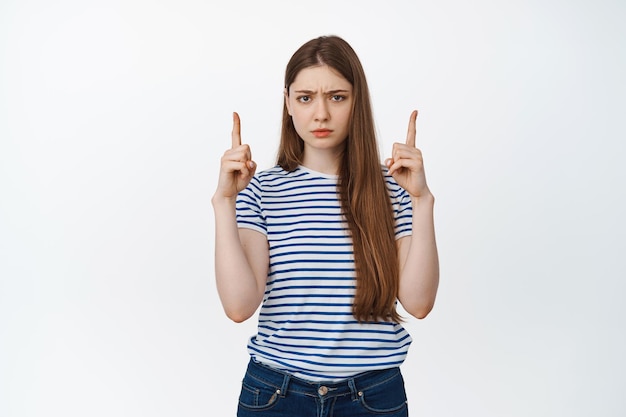 Upset gloomy girl frowning, pointing fingers up at empty space, standing in casual clothes against white background