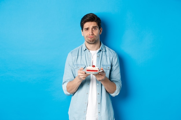 Upset and gloomy man holding birthday cake, sulking sad, standing against blue background.