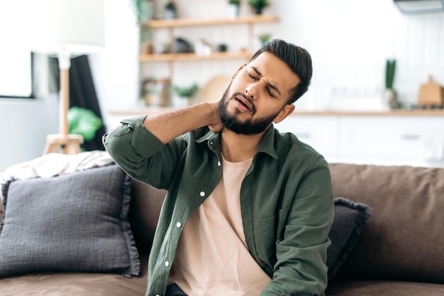 Upset indian or arabian young man sitting on sofa in cozy\
living room massaging his neck tired after a hard day at work\
suffering from joint problems pinched nerve in neck need rest