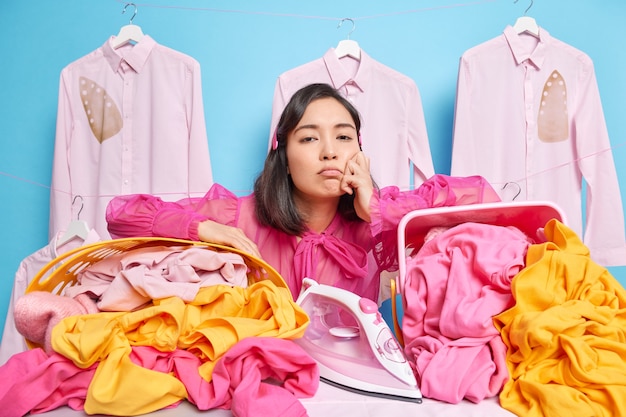 Upset laundry worker leans at baskets full of linen feels tired busy ironing poses against blue wall