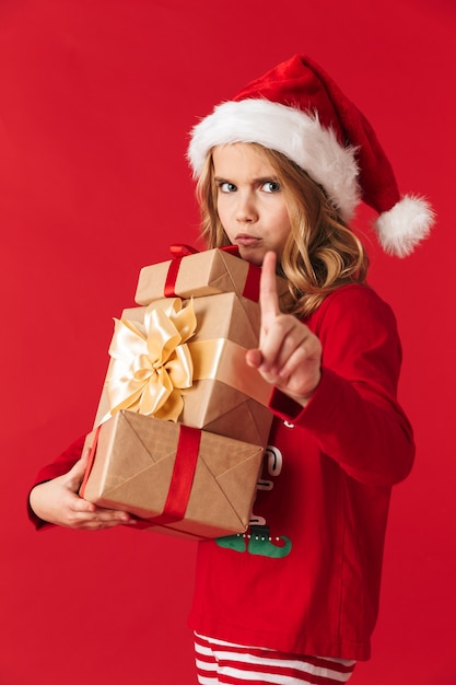 Upset little girl wearing Christmas costume standing isolated, holding gift boxes