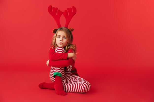 Upset little girl wearing Christmas raindeer costume sitting isolated