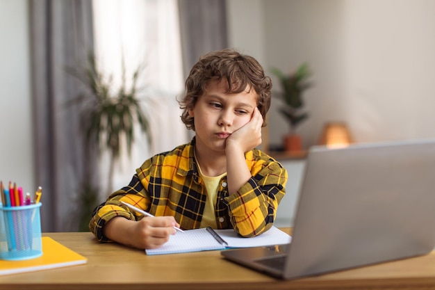 Upset little schoolboy watching uninteresting online lesson on laptop feeling bored at home empty space