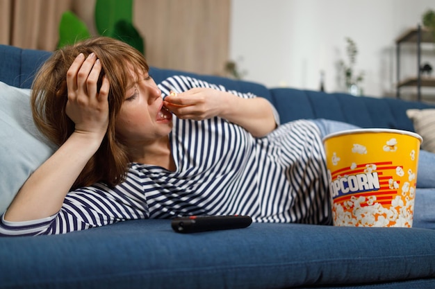 Upset lonely woman liyng on sofa eating popcorn and watching boring tv