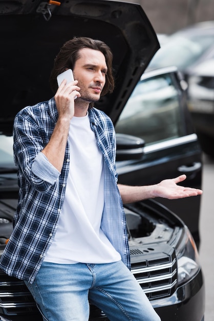 Upset man in casual wear talking on smartphone while standing near broken auto with open trunk car