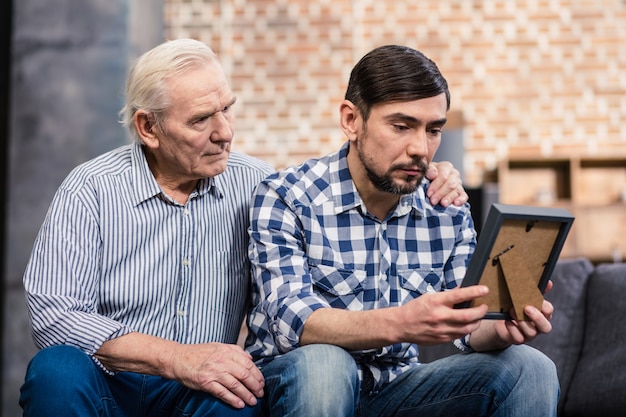 upset man holding a photo frame while his father supporting him at home