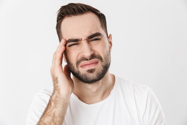 Upset man wearing blank t-shirt standing isolated over white wall, suffering from a strong migraine