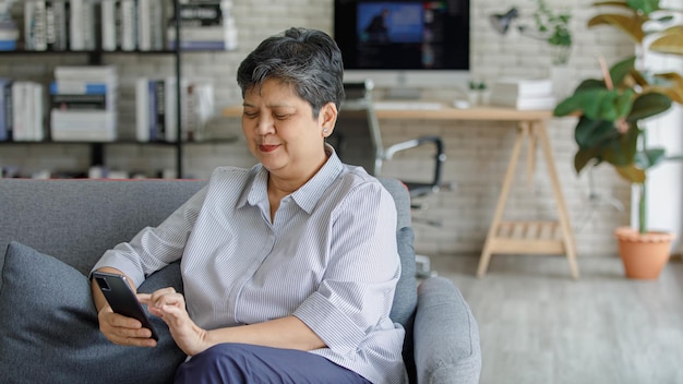 Upset mature Asian woman frowning and browsing smartphone while sitting on sofa in modern living room at home
