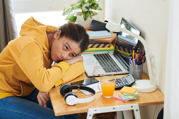 Upset mixed race student girl with Afro hair leaning head on crossed arms on table with laptop and headphones and looking at lamp