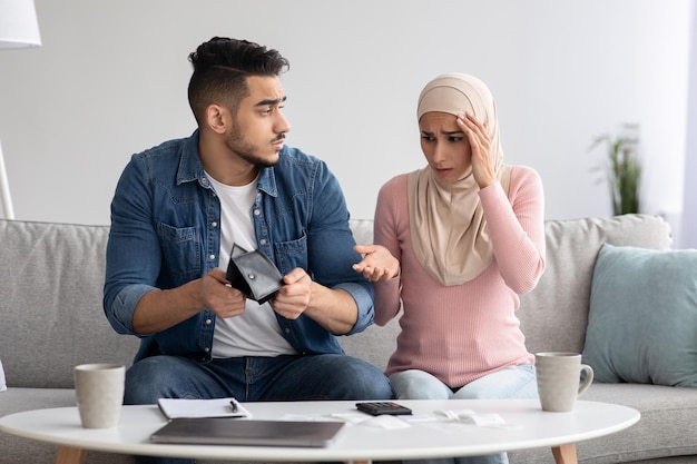 Upset muslim family with empty wallet counting their spendings\
together, sitting on sofa in living room at home. shocked young\
arab man and woman working on family budget, financial hangover\
cocnept